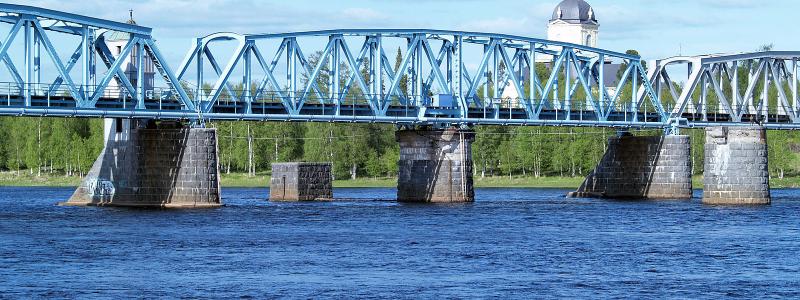 Järnvägsbron över Torne-älv. En bro mellan svenska Haparanda och den östra delen av den finska staden Torneå. Ursprungligen kunde mittspannet vändas för att låta höga skepp passera. Bakom ryttarna kyrkan i Alatornio. Foto: Wikipedia, kredit: Bobo11